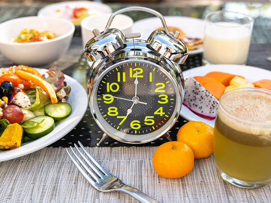 Una sveglia analogica argentata è posta su un tavolo tra vari prodotti per la colazione, tra cui insalata, frutta, succo di frutta e un bicchiere di latte.