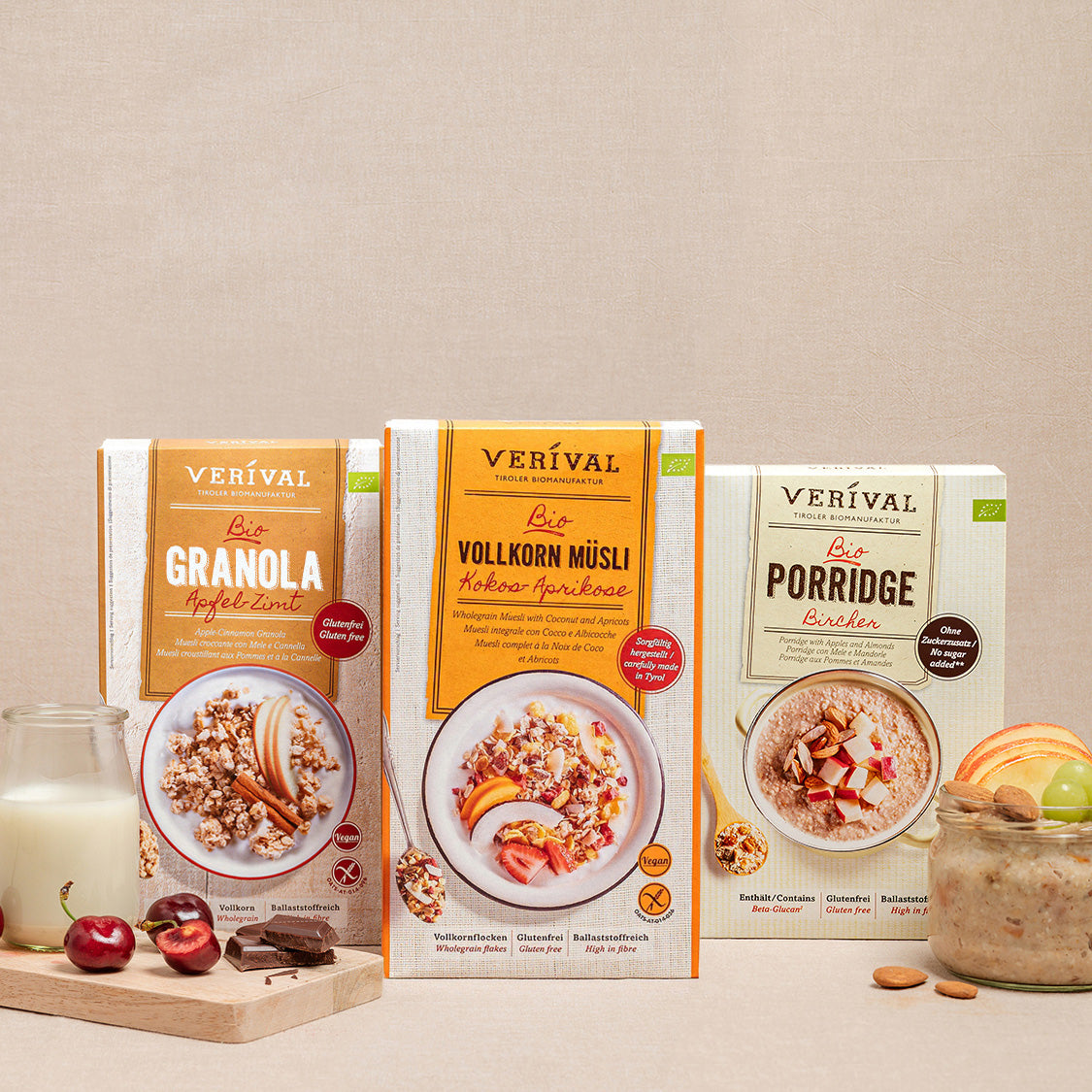 Drei Müslischachteln mit den Aufschriften Crunchy, Vollkorn Müsli und Porridge werden mit einem Glas, Kirschen und Mandeln auf beigem Hintergrund präsentiert.