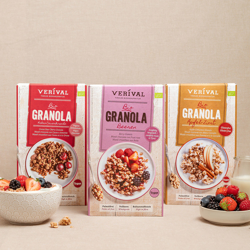 Trois boîtes de granola Verival aux saveurs variées se tiennent debout sur une surface beige, avec un bol de granola et de baies mélangées devant elles.