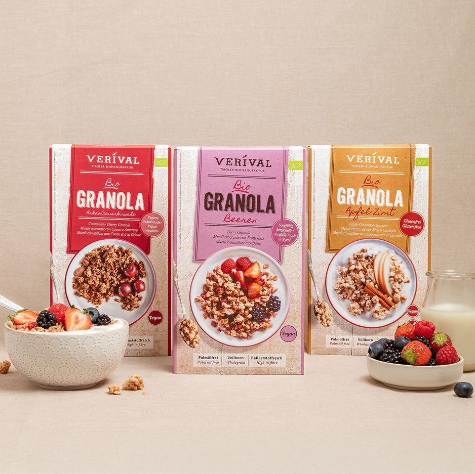 Trois boîtes de muesli complet Verival aux saveurs différentes sont présentées avec un bol de muesli, un verre de lait et diverses baies sur un fond beige.