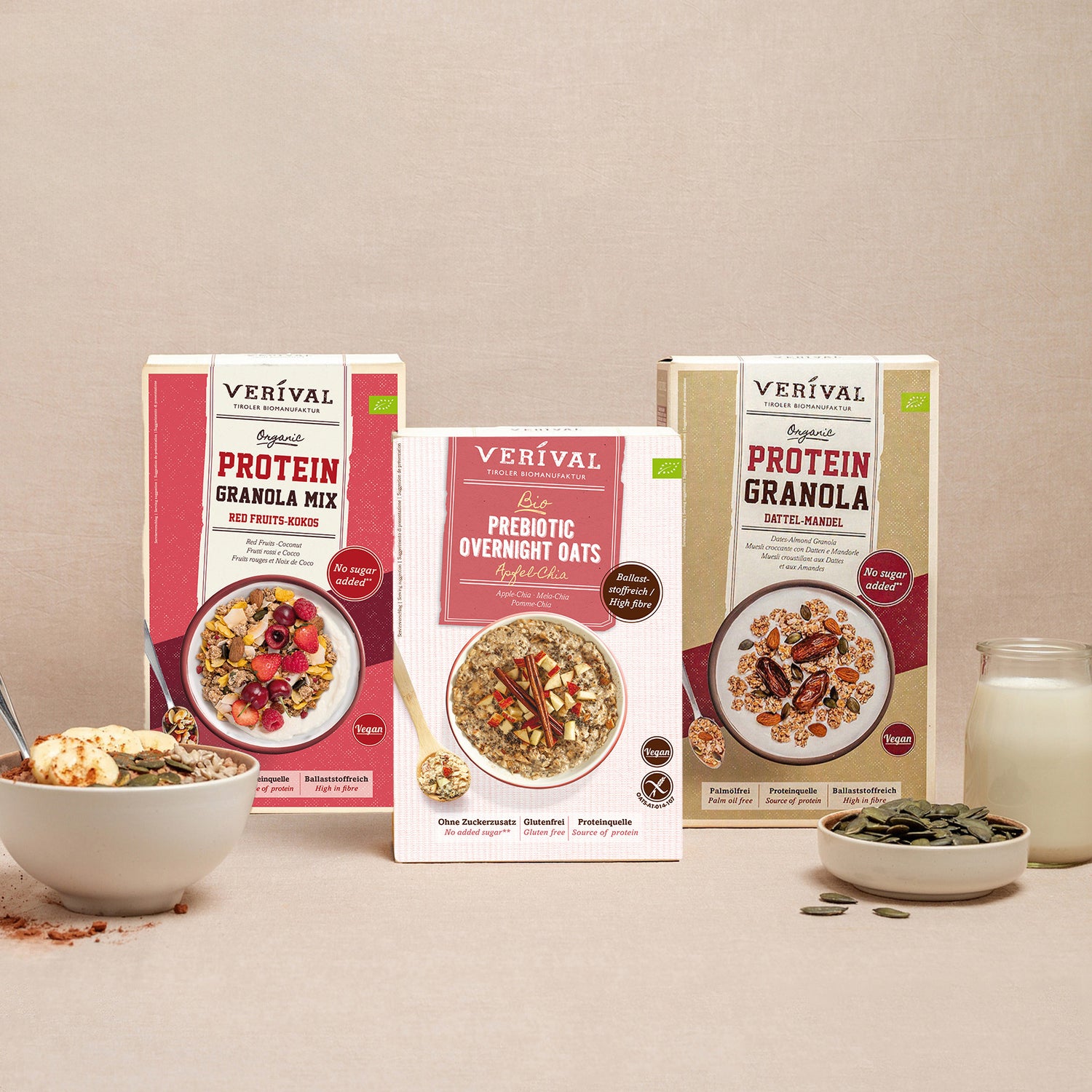 Trois paquets de céréales pour petit-déjeuner Verival, comprenant un mélange granola protéiné, des flocons d'avoine prébiotiques pour la nuit et un granola protéiné. À l'avant, un bol de muesli, une cuillère, des noix et un verre de lait.