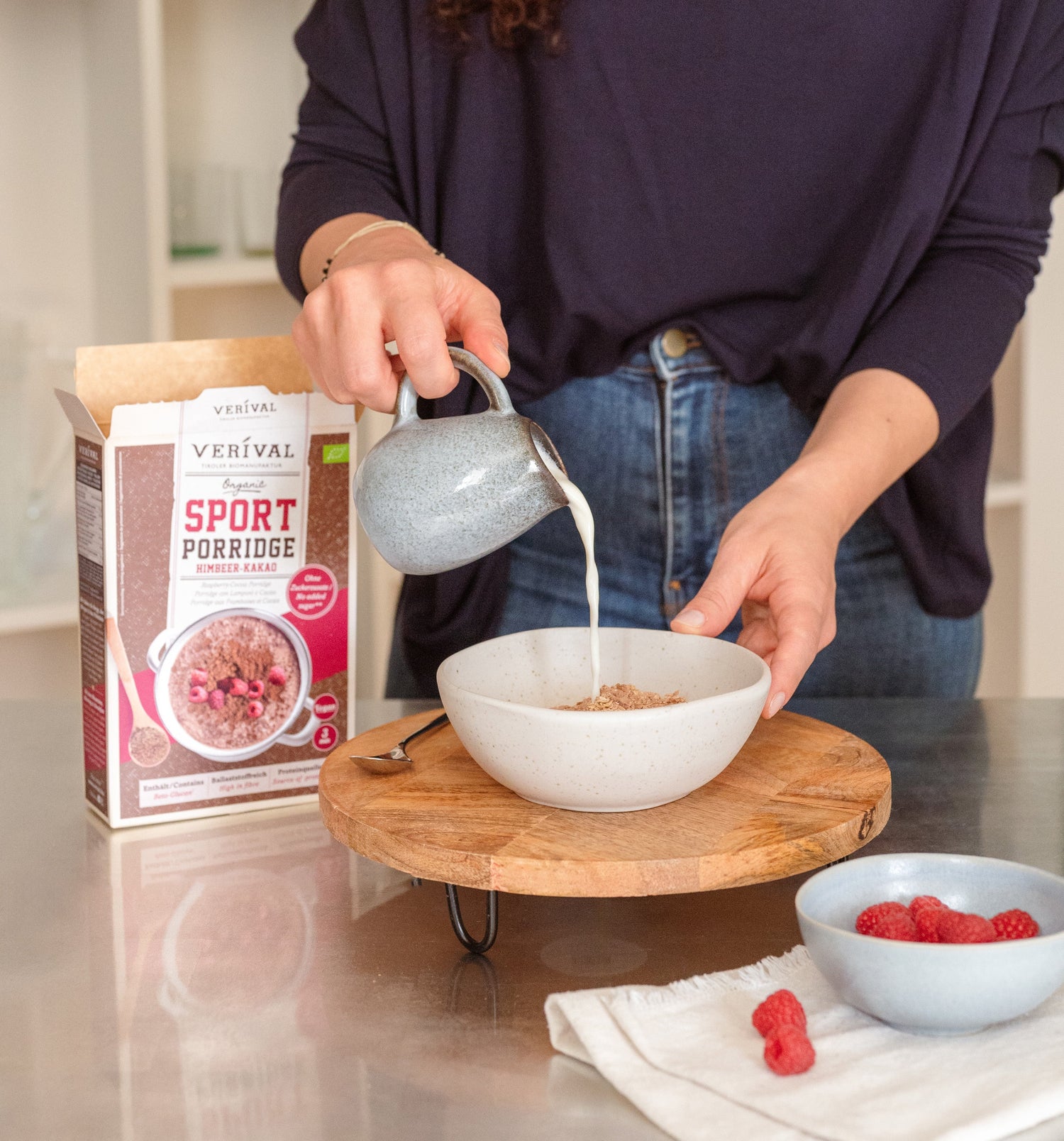 Une personne verse du lait dans un bol de porridge à côté d'une boîte étiquetée Sport Porridge et d'un petit bol de framboises sur un plateau en bois.