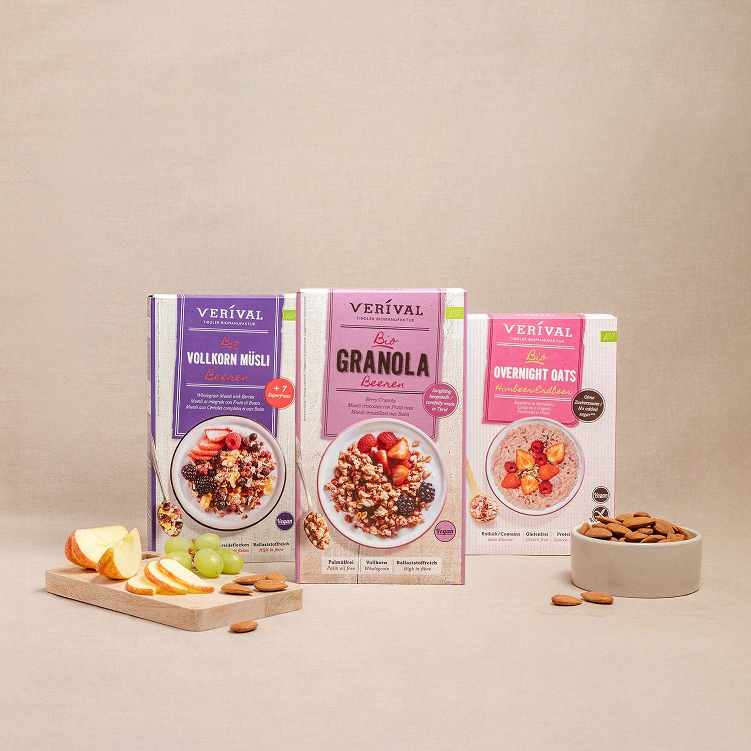 Trois paquets de muesli Verival (muesli complet, granola, flocons d'avoine de nuit) sont présentés avec un bol d'amandes et une planche à découper avec des tranches de pomme et de raisin.