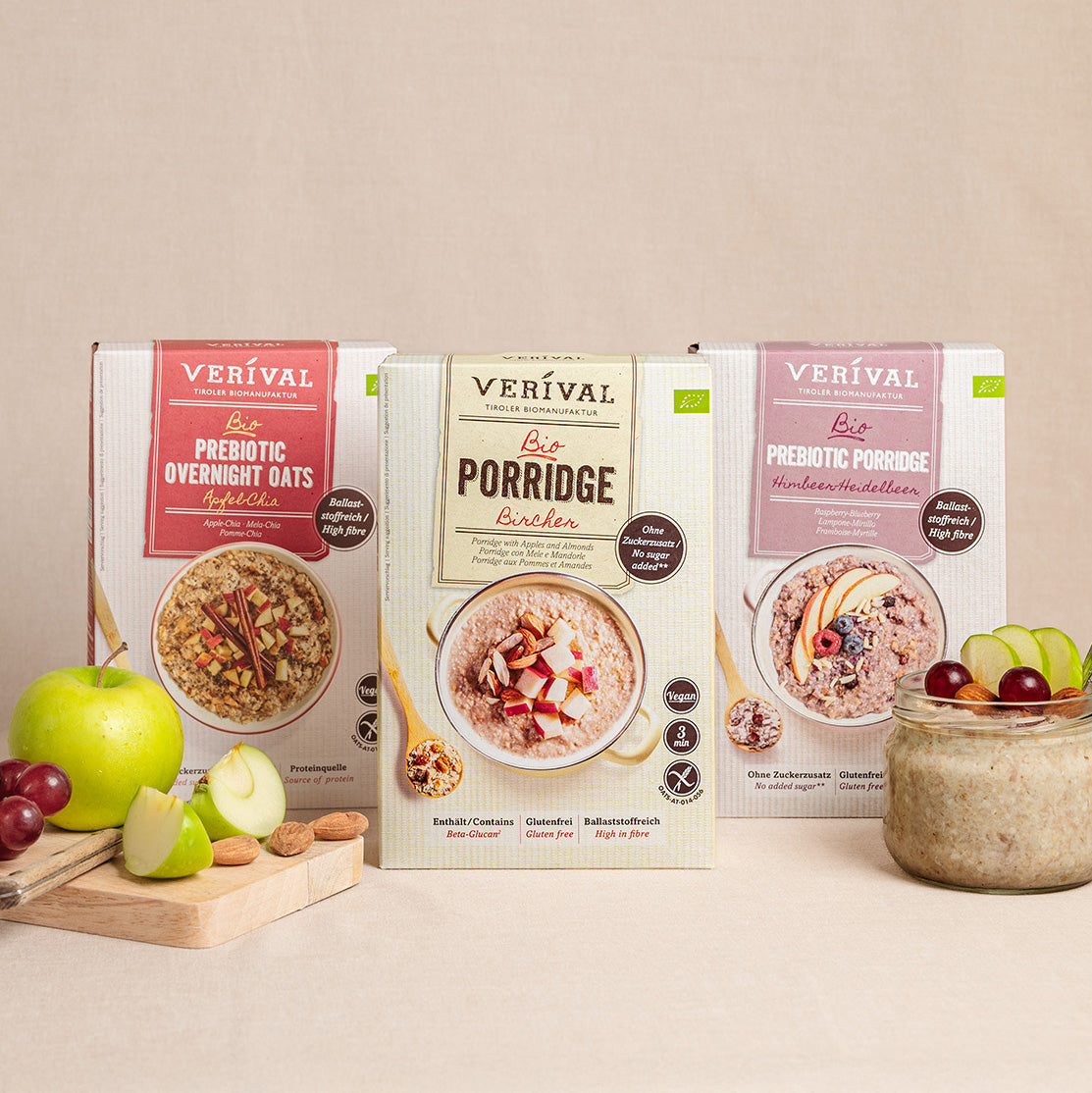 Trois paquets de céréales pour petit-déjeuner Verival – flocons d'avoine prébiotiques pour la nuit, porridge biologique et porridge prébiotique – présentés avec un bol de porridge, des tranches de pomme et des raisins à l'avant.