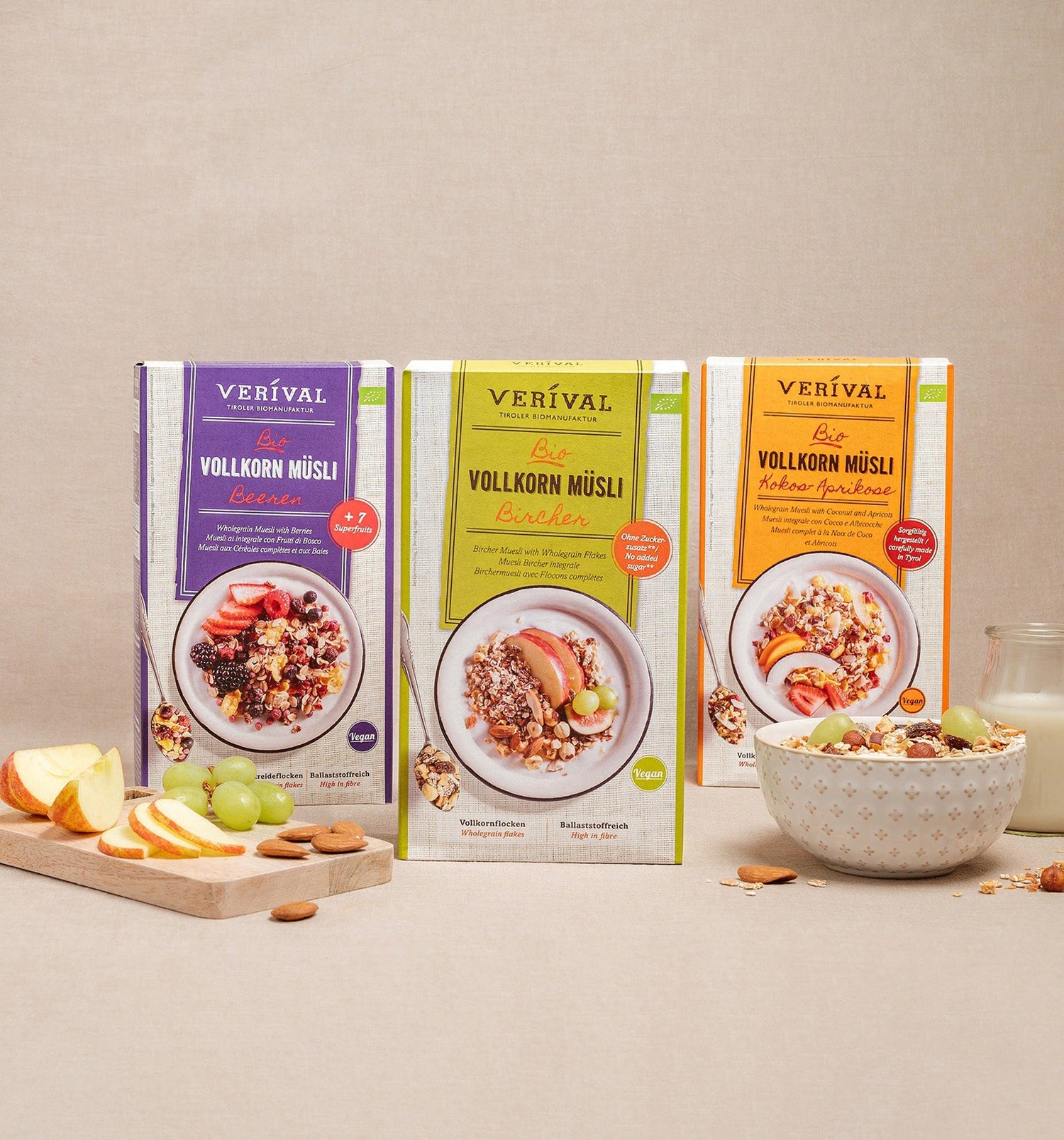 Trois boîtes de muesli complet bio Verival sont dressées, devant elles un bol de muesli, des tranches de pomme, des raisins, une cuillère et un verre de lait.