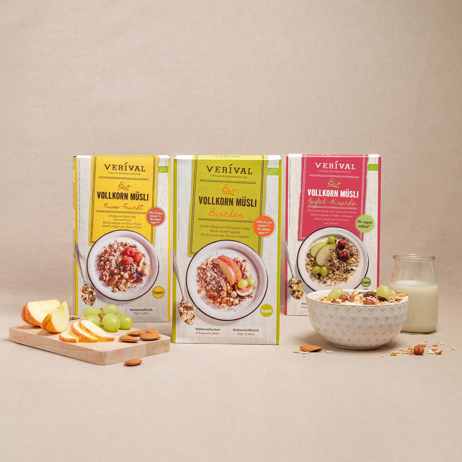Trois boîtes de muesli complet Verival aux saveurs variées sont présentées debout avec un bol de muesli, un verre de lait, des tranches de pomme et des raisins devant.