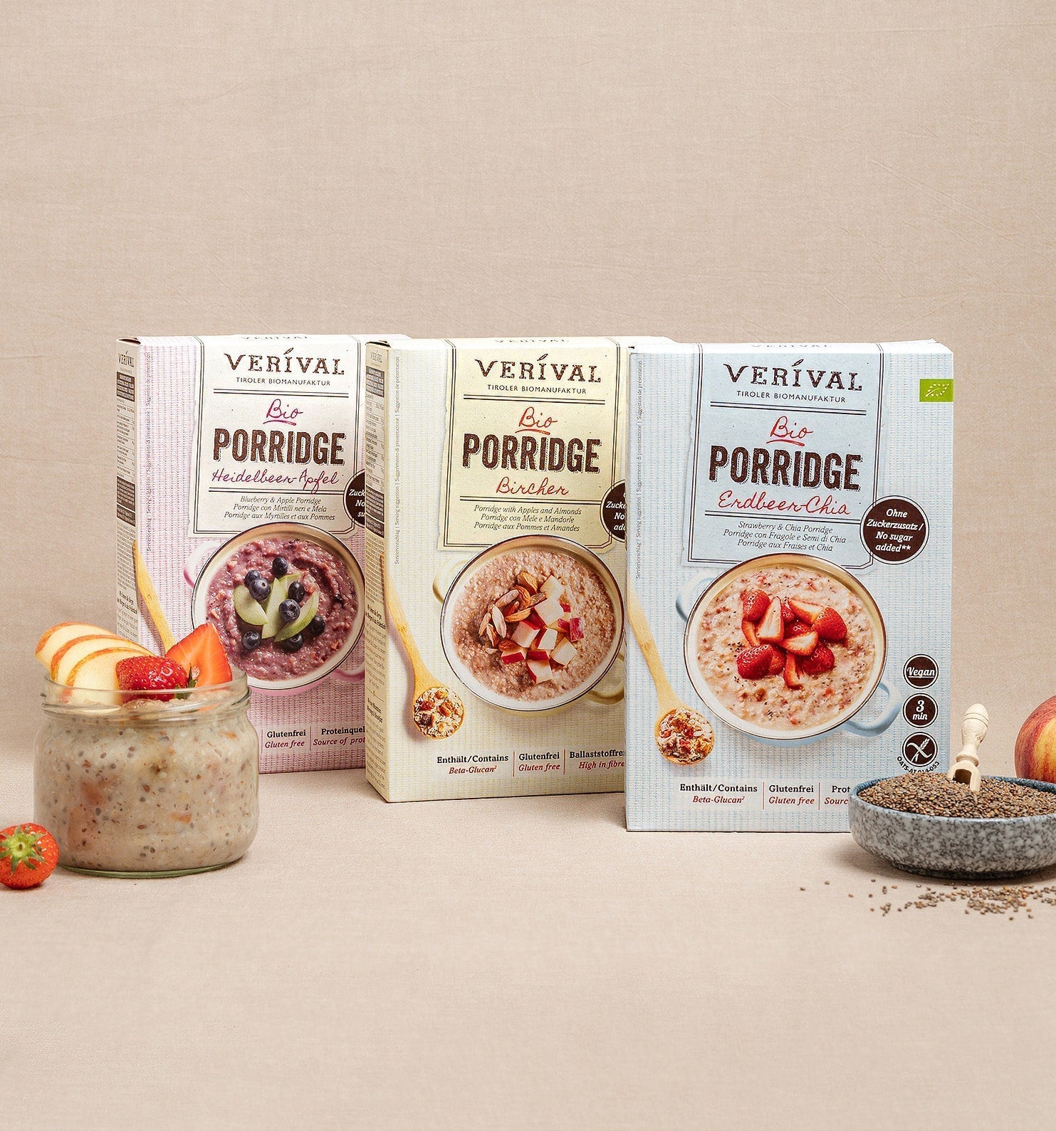 Trois boîtes de porridge biologique Verival (variétés pomme, Bircher et chia) sont présentées avec un verre de porridge, des fruits frais, des graines de chia et une pomme sur fond beige.