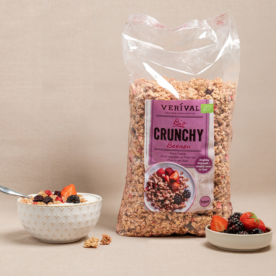 Eine Tüte Verival Bio Crunchy Beeren-Müsli steht neben einer Schale Müsli mit Milch und verschiedenen Beeren, und auf dem Tisch stehen zusätzliche Beeren.