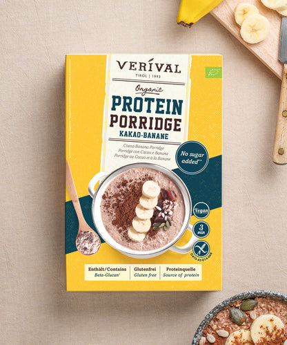 Eine Packung Verival Bio Bio Kakao-Banane Protein Porridge neben einer Schüssel mit dem zubereiteten Haferbrei, garniert mit Bananenscheiben und Kakaopulver.