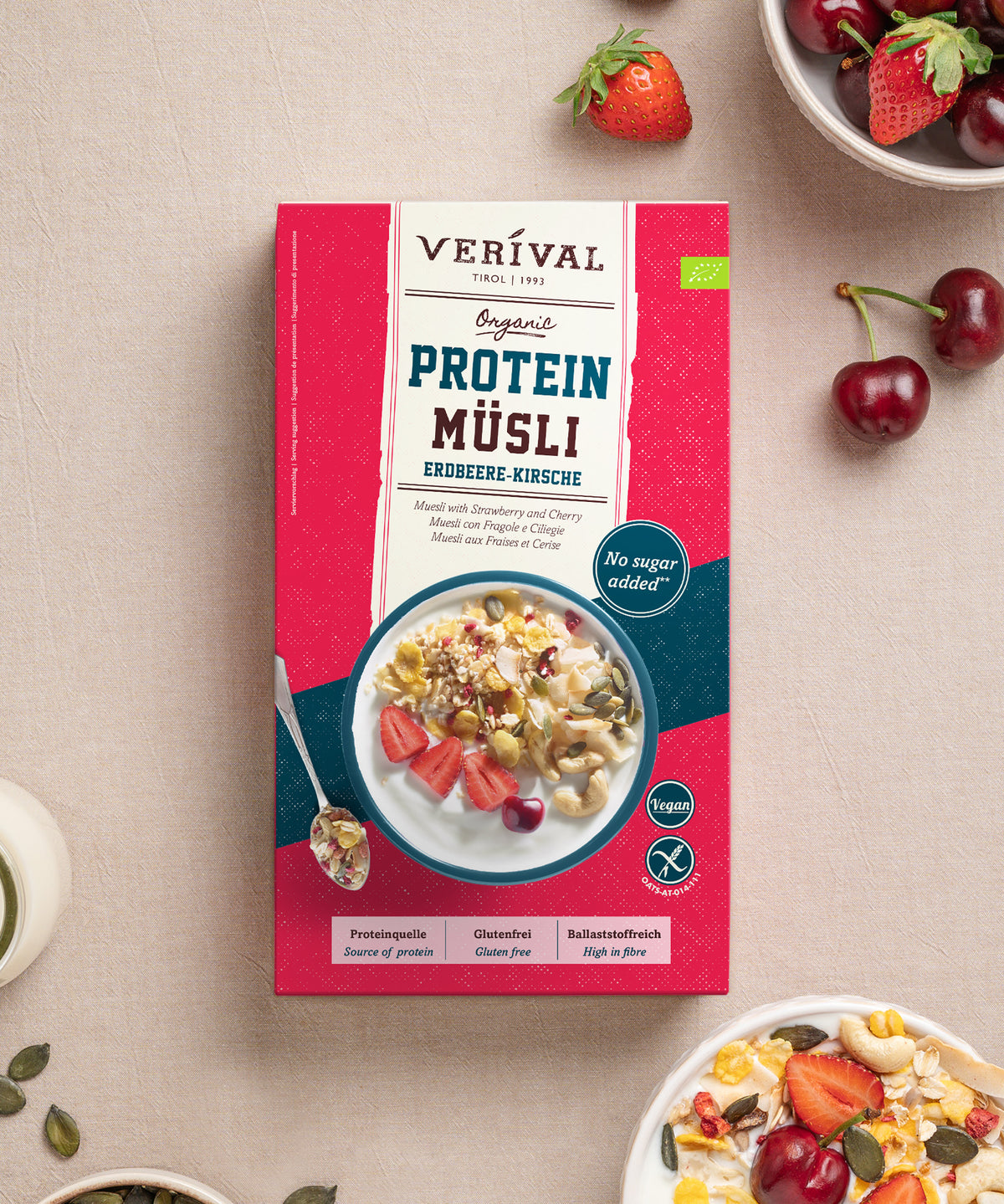 Eine Packung Bio Erdbeere-Kirsche Protein Müsli von Verival Bio GmbH als pflanzliche Proteinquelle, abgebildet zusammen mit losen Kirschen, einer Schüssel Müsli und einem Löffel.