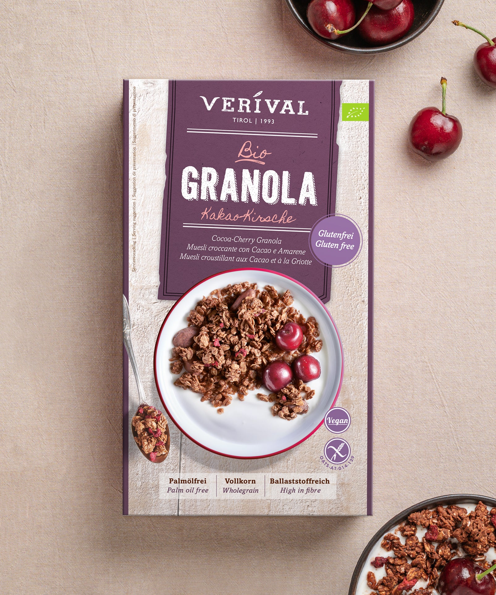 Eine Schachtel Verival Bio GmbH Bio Kakao-Kirsche Granola glutenfrei steht auf einer hellen Fläche, umgeben von Kirschen und einer Schale veganes, glutenfreies Knuspermüsli mit Kirschen.