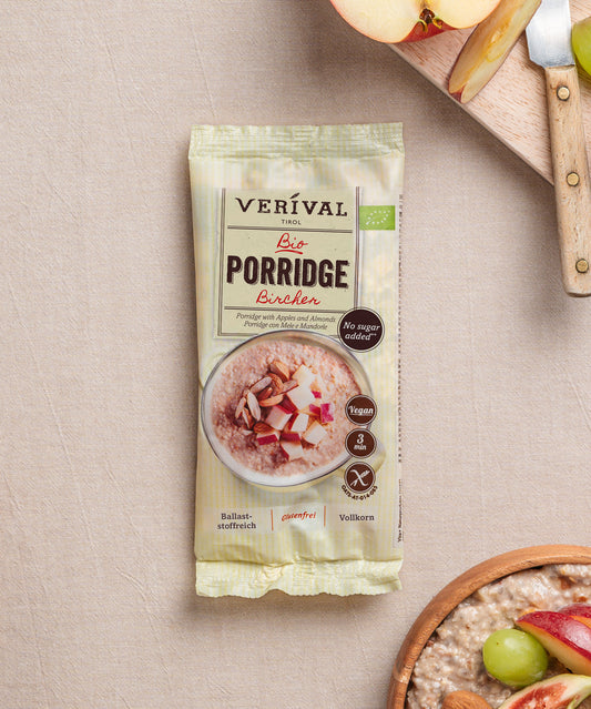 Eine Packung Erdbeer-Chia-Porridge der Verival Bio GmbH liegt auf einer beigen Oberfläche neben einem geschnittenen Apfel, einem Holzmesser, Weintrauben und einer Schüssel pflanzlichem Frühstücksbrei mit Früchten.