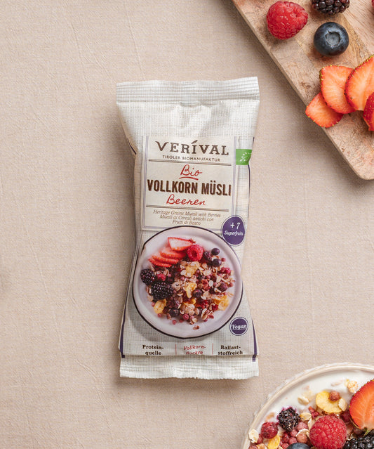 Eine Einzelportionspackung Beeren Vollkorn Müsli von Verival Bio steht auf einem Tisch neben einem mit Früchten belegten Holzbrett und einer Schüssel ballaststoffreichem Müsli.