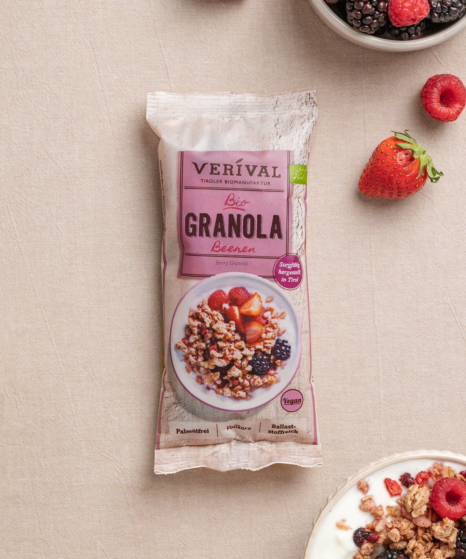 Eine Einzelportions-Packung Beeren Granola von Verival Bio liegt auf einem beigen Untergrund neben einer Joghurtschale mit knusprigem Granola und leuchtenden Erdbeeren, Himbeeren und Brombeeren. 