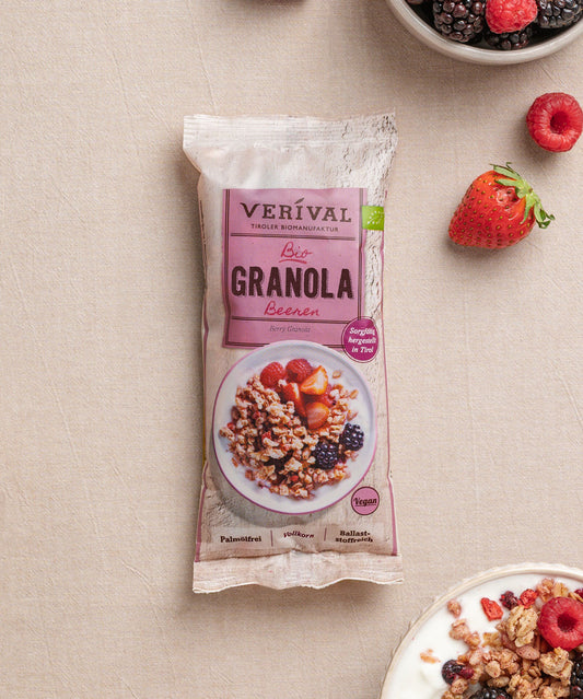 Eine Einzelportions-Packung Beeren Granola von Verival Bio liegt auf einem beigen Untergrund neben einer Joghurtschale mit knusprigem Granola und leuchtenden Erdbeeren, Himbeeren und Brombeeren. 