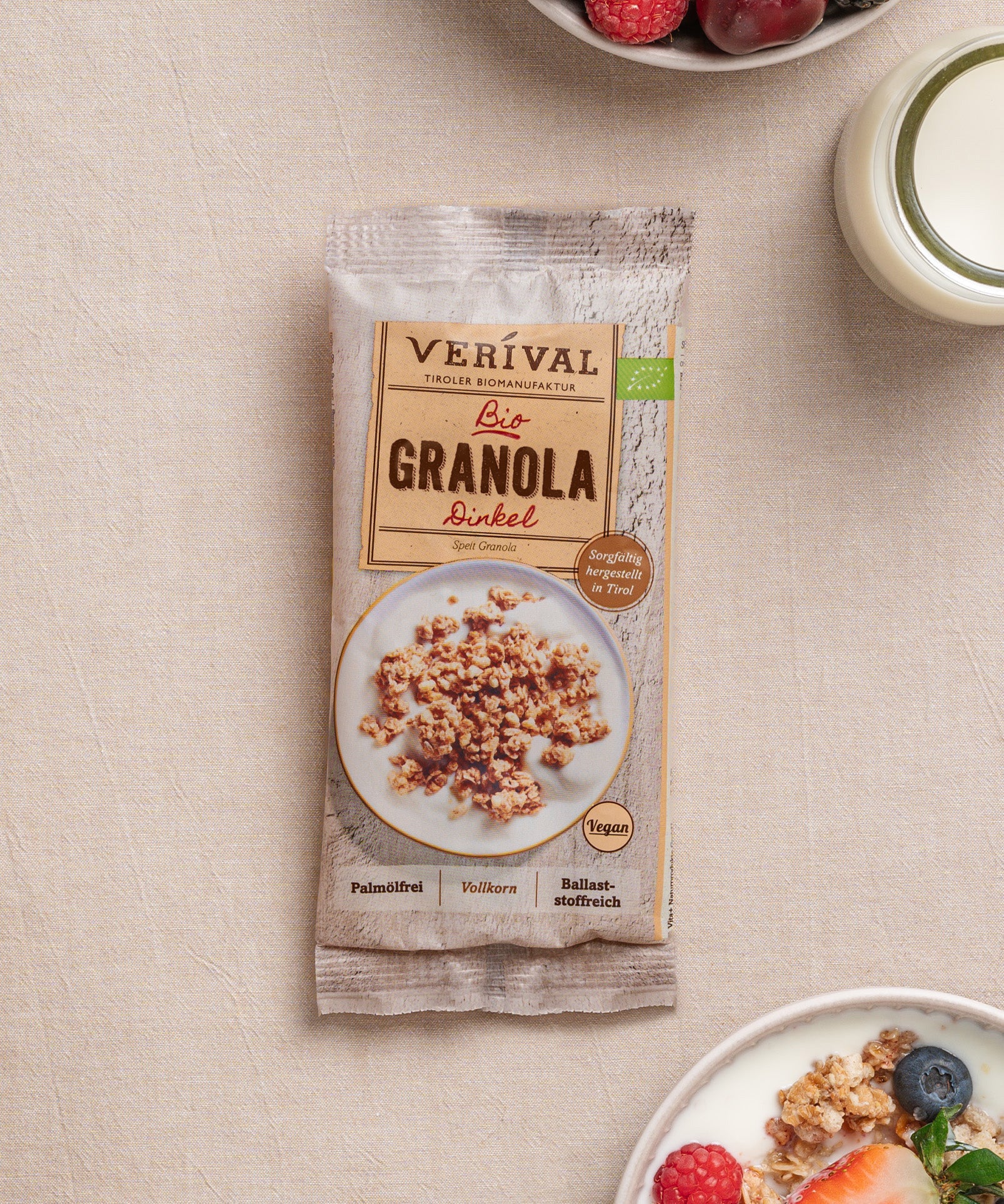 Eine Packung Dinkel-Granola von Verival Bio liegt auf beigem Untergrund, neben einer Schüssel des knusprigen veganen Granolas, serviert mit Milch und Beeren. Dieses ballaststoffreiche Frühstück ist der ideale Start in den Tag.