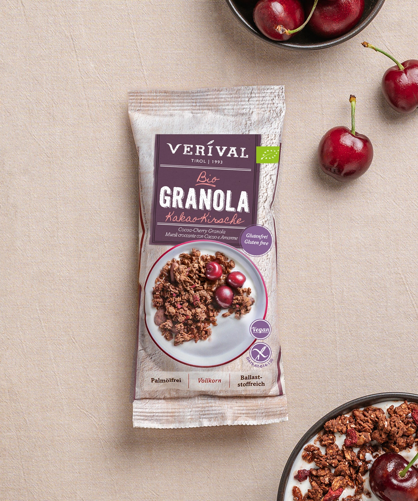 Eine Packung Verival Bio GmbH Bio Kakao-Kirsche Granola glutenfrei, eine vegane und ballaststoffreiche Leckerei, steht auf einem Tisch mit frischen Kirschen und Schalen mit glutenfreiem Knuspermüsli und Kirschen daneben.