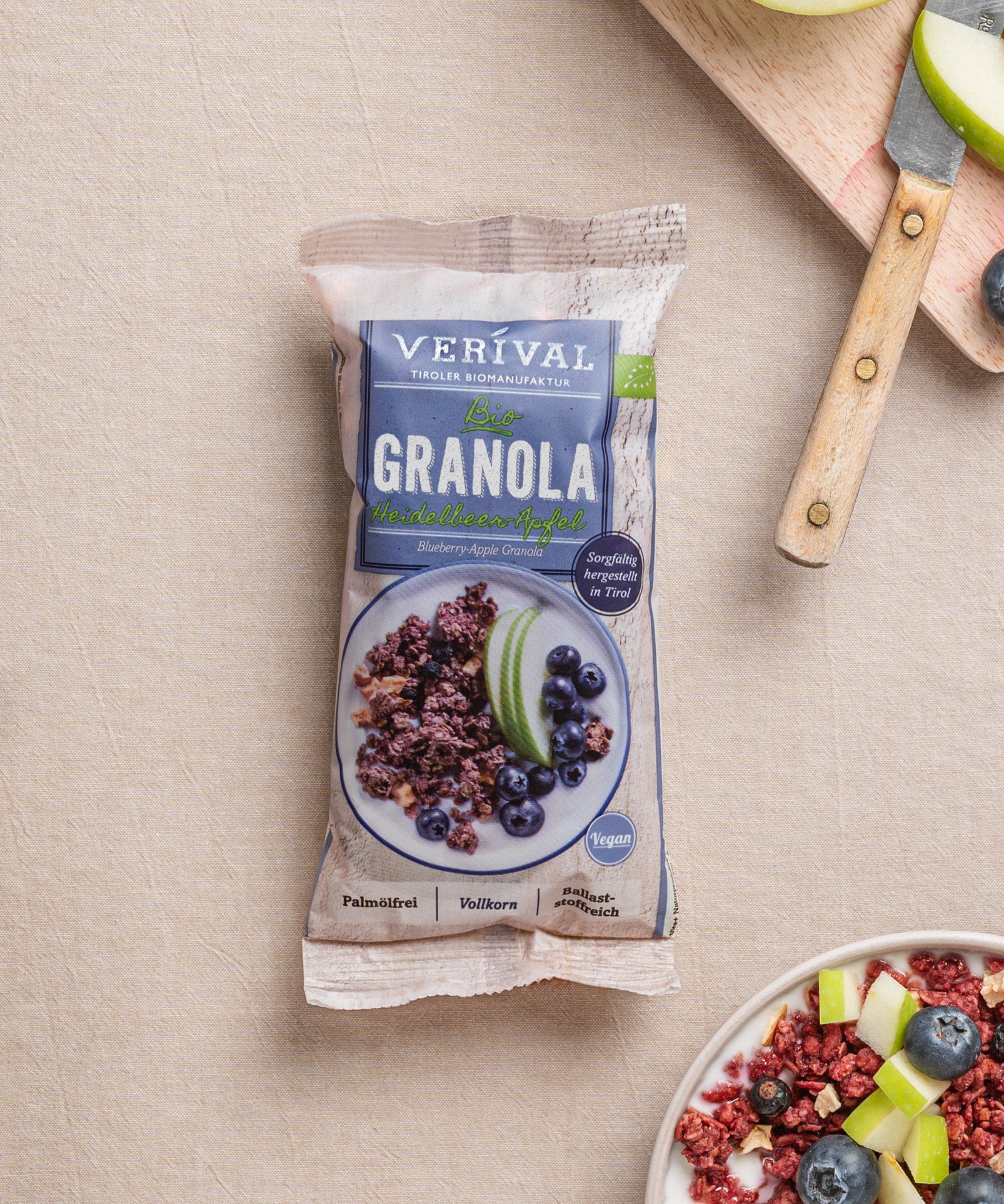Eine Packung Heidelbeer-Apfel-Granola von Verival Bio steht auf einer beigen Fläche, daneben ein Holzbrett mit geschnittenen Äpfeln, Heidelbeeren und Granola. 