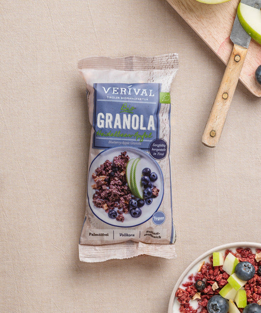 Eine Packung Heidelbeer-Apfel-Granola von Verival Bio steht auf einer beigen Fläche, daneben ein Holzbrett mit geschnittenen Äpfeln, Heidelbeeren und Granola. 