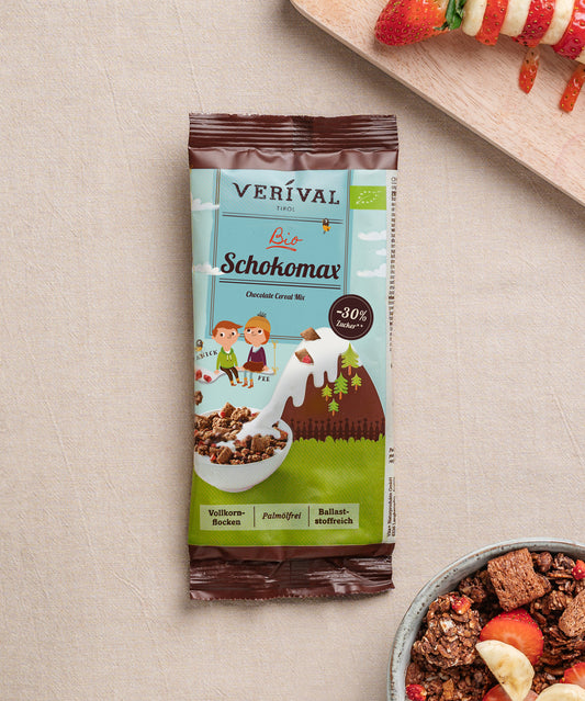 Auf einem beigen Untergrund liegt ein Bio Schokomax Schokoriegel der Verival Bio GmbH neben einer Schale Knuspermüsli mit Früchten und einem teilweise abgebildeten Teller mit Fruchtspießen.