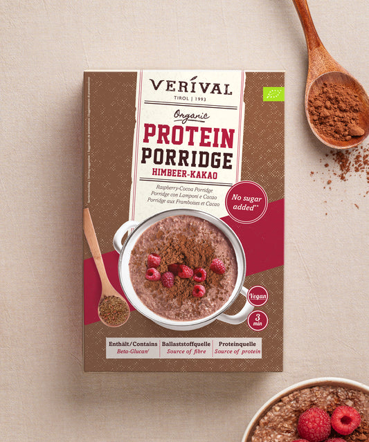 Eine rechteckige Verpackung von Verival Bio Himbeer-Kakao Protein Porridge neben einer Schüssel mit fertig zubereitetem Porridge, garniert mit Himbeeren und Kakaopulver, daneben liegen Holzlöffel.