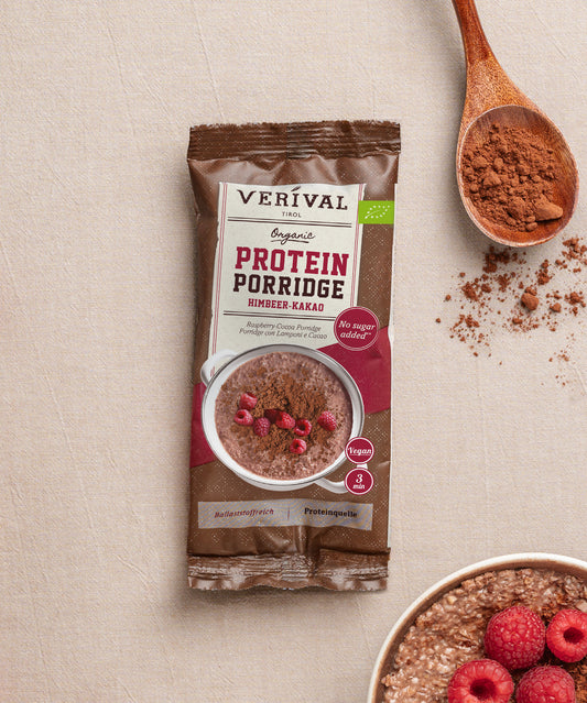 Eine Packung Bio Protein Porridge Himbeer-Kakao der Verival Bio GmbH liegt auf einer beigen Oberfläche neben einem Löffel Kakaopulver, frischen Himbeeren und einer Schüssel ballaststoffreichem veganen Frühstück.