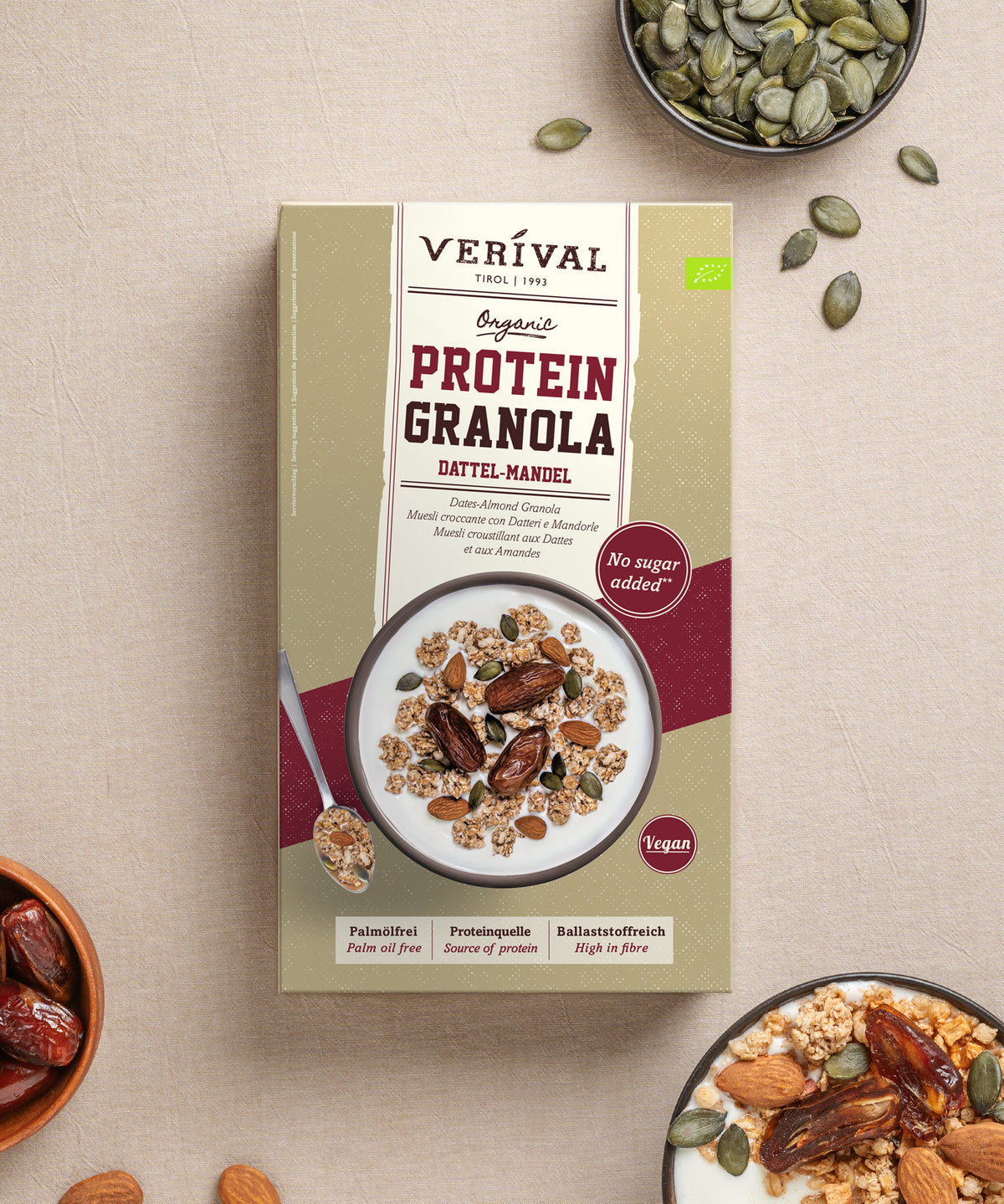 Packung Verival Bio Bio Dattel-Mandel Protein Granola, abgebildet mit einer Schüssel Granola, garniert mit Nüssen und Datteln, einer kleinen Schüssel mit Kürbiskernen und einer Schüssel mit ganzen Datteln.