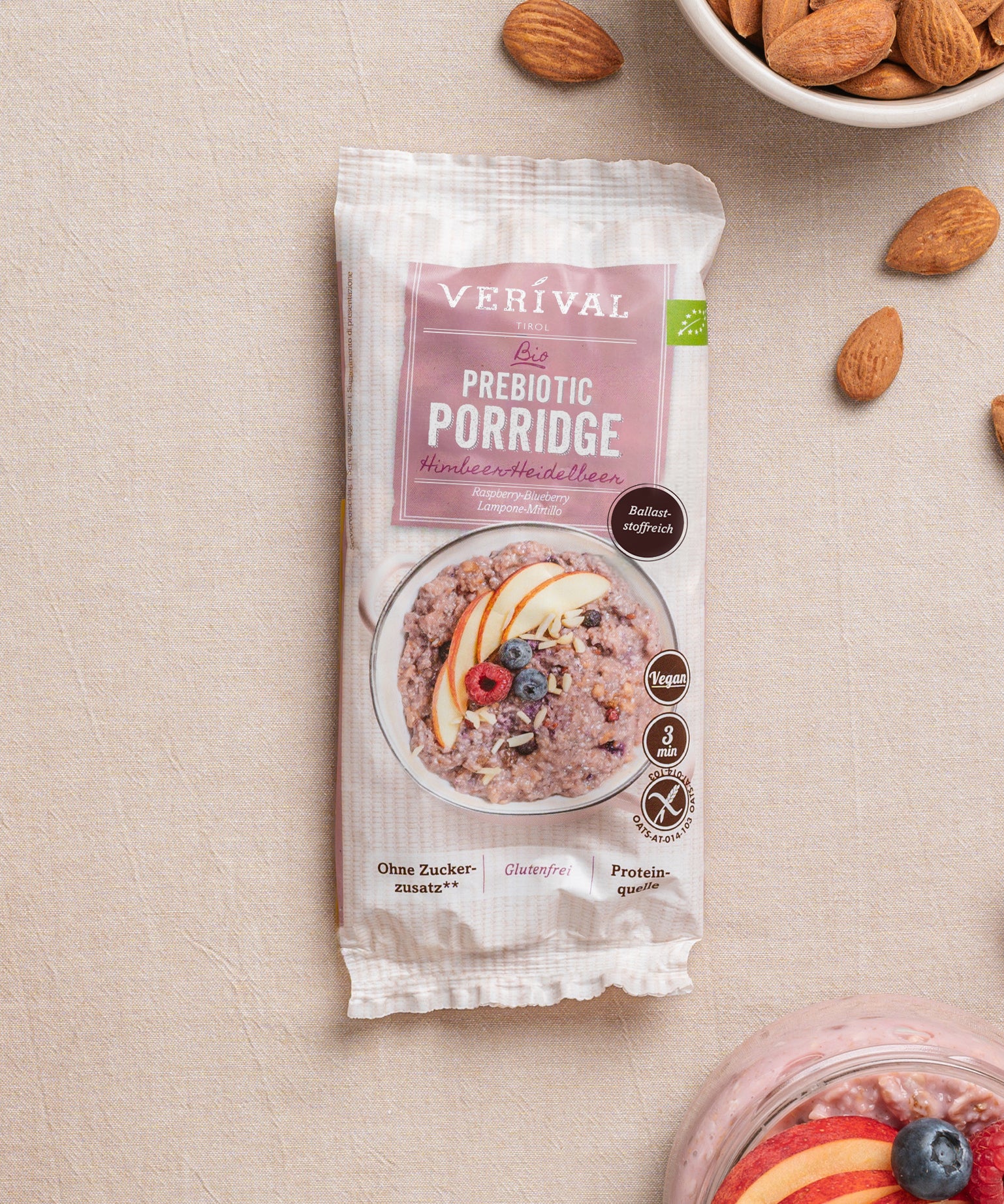 Eine Packung Erdbeer-Chia-Porridge der Verival Bio GmbH, gekennzeichnet als vegan, glutenfrei und proteinreich, steht auf einer beigen Fläche neben Mandeln und einer Schüssel Pflanzlich Frühstück-Porridge.