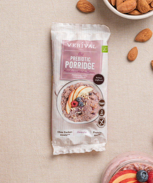 Eine Packung Erdbeer-Chia-Porridge der Verival Bio GmbH, gekennzeichnet als vegan, glutenfrei und proteinreich, steht auf einer beigen Fläche neben Mandeln und einer Schüssel Pflanzlich Frühstück-Porridge.