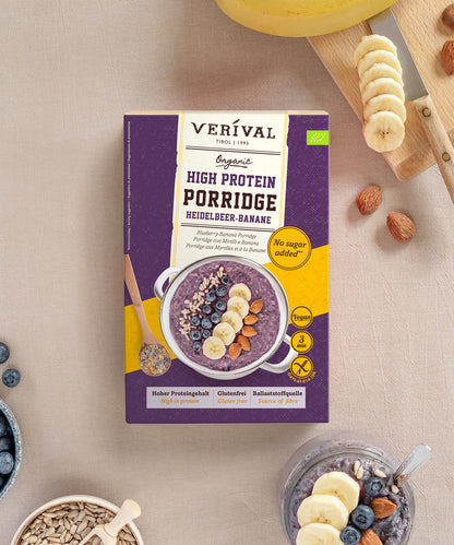 Eine Packung Verival Bio Heidelbeere-Banane High Protein Porridge liegt über einer bereits angerichteten Schüssel, umgeben von Bananenscheiben und Mandeln.