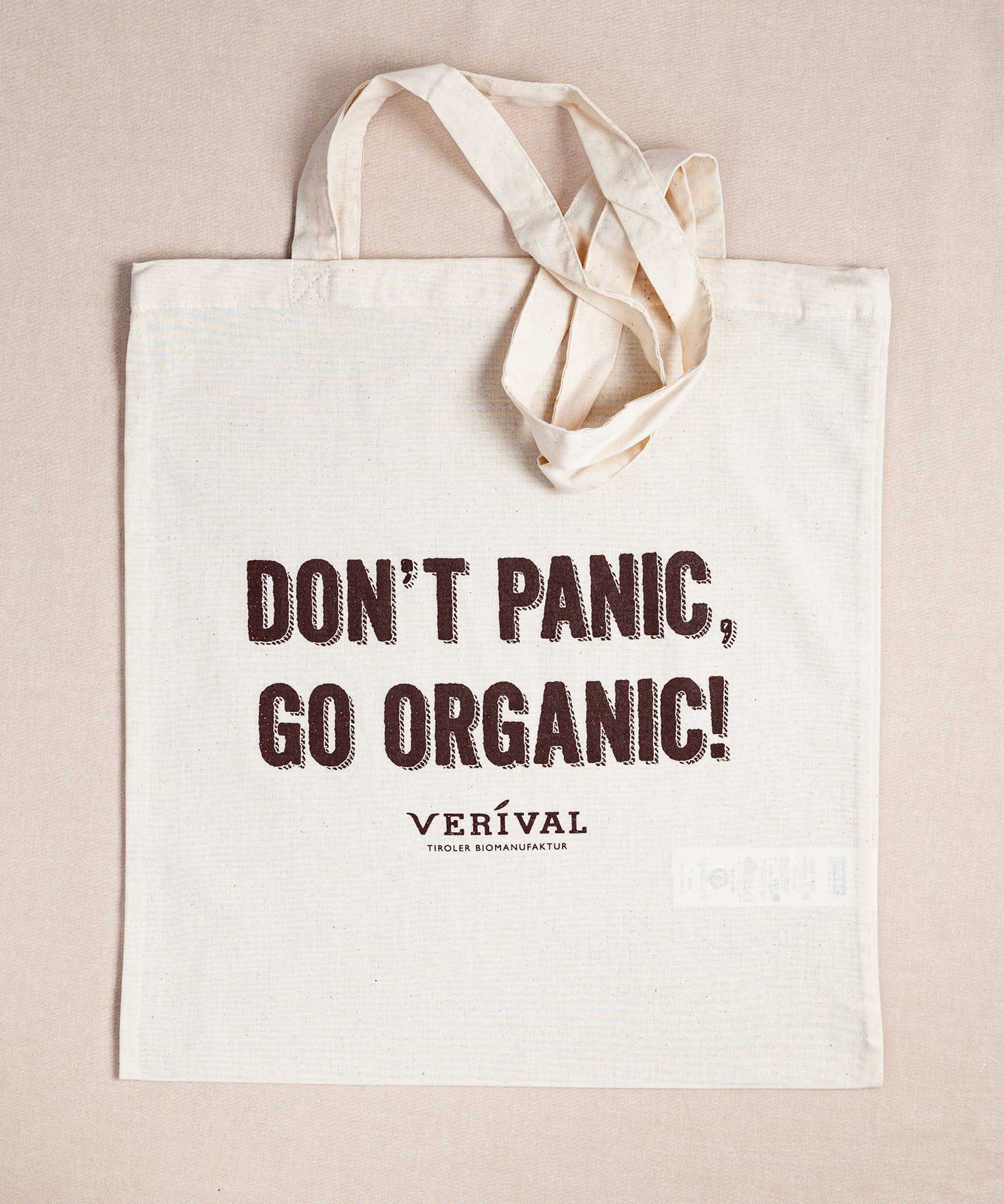 Die Verival Bio Stofftasche von Verival Bio ist eine cremefarbene Tragetasche aus strapazierfähiger Bio-Baumwolle. Sie hat lange Henkel und trägt den auffälligen Slogan „Don't Panic, Go Organic!“ mit dem Markennamen VERIVAL darunter – perfekt zum Einkaufen.