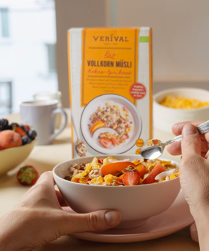 Eine Person hält einen Löffel über eine Schüssel mit „Verival Bio Kokos-Aprikose Vollkorn-Müsli“ mit Erdbeeren und Milch. Im Hintergrund sind die Müsli-Packung, die Obstschale, die Tasse und Cornflakes auf einem hellen Tisch zu sehen. Dieses Frühstück ist ballaststoffreich und gesund.