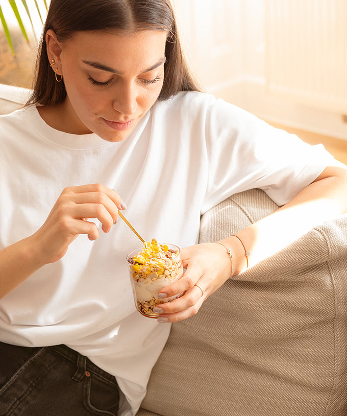 Eine Frau in einem weißen T-Shirt und mit goldenem Schmuck entspannt sich auf dem Sofa und genießt das glutenfreie, ballaststoffreiche Bio-Kokos-Aprikosen-Vollkorn-Müsli der Verival Bio GmbH mit Joghurt und Obst.