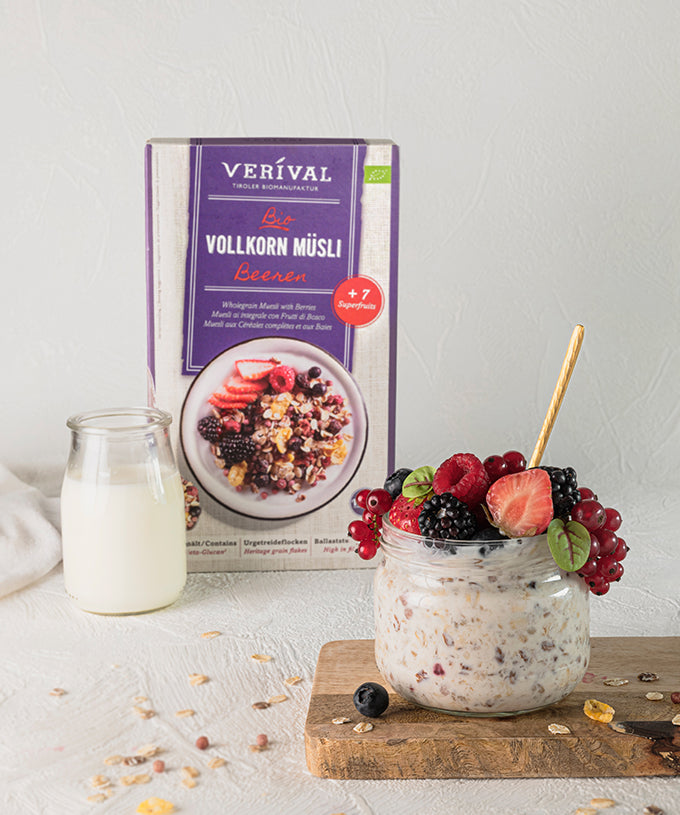 Ein Glas veganes Müsli mit Beeren steht auf einem Holzbrett neben einem Glas Milch und einer Packung „Bio-Beeren-Vollkorn-Müsli“ der Verival Bio GmbH. Superfrüchte und Müslistückchen sind auf der weißen Oberfläche verstreut.